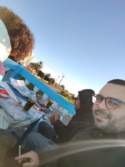 Three individuals gather outdoors, likely at a park, under a clear blue sky. A table is set with glasses of a dark beverage, possibly tea, and other small items. Two individuals are seated, one wearing glasses and smiling, holding a cigarette. Another with a hoodie on. A third person with large curly hair is partially visible. The background includes trees, a power pole, and a road.