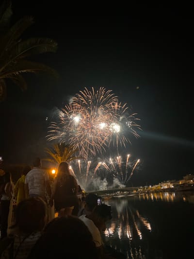 The image depicts a nighttime scene of a fireworks display over the Gilão River in Tavira, Portugal. In the foreground, a crowd of people are gathered on the riverbank, silhouetted against the bright explosions in the sky. Palm trees are visible on the left side of the image, with their fronds casting shadows. The fireworks are orange and white, with some trails of smoke rising into the dark sky. The lights from the fireworks are reflected in the dark water of the river, creating a shimmering effect. In the background, buildings and a bridge are illuminated by the fireworks, suggesting a populated area along the river.