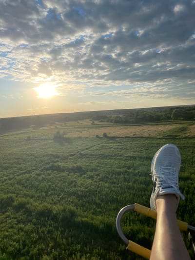 The image captures a first-person perspective from a hot air balloon ascending over a rural landscape. In the foreground, a person's right leg and foot, clad in a white athletic shoe, are visible, resting on a yellow handle of the balloon's basket. The leg is illuminated by sunlight, suggesting a warm day. The background reveals a sprawling green field dotted with trees and some patches of drier vegetation, indicative of farmland. A distant tree line forms the horizon under a sky filled with a mixture of white and grey clouds. The sun is partially obscured by the clouds, casting a warm, golden light across the scene. The geographical location is specified as Турово, Russia.