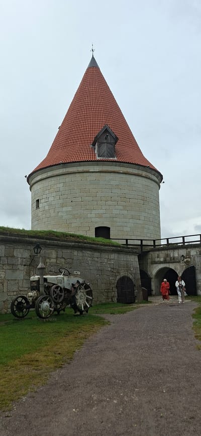 The image shows the Kuressaare Episcopal Castle in Kuressaare, Estonia. The castle is a well-preserved medieval fortress with a distinctive conical red-tiled roof and stone walls. In the foreground, an old, rusty tractor is parked on the grass. The tractor is a vintage model with large spoked wheels. To the right of the tractor, a stone wall surrounds the castle grounds, with arched gateways leading inside. Two women are walking through one of the archways, dressed in casual attire. The sky is overcast, suggesting a cloudy day. The overall scene depicts a historical landmark with elements of both the past and present.