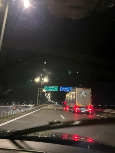 A nighttime shot from a vehicle on a highway near Mikrevo, Bulgaria. The vehicle is approaching a road sign indicating that going straight leads to Sofia, while taking a left turn leads to Strumyani. Ahead, there is a large truck. The road is illuminated by streetlights. The image appears to have been taken from inside the car, potentially the passenger seat.