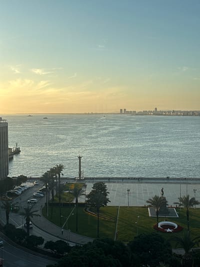 The image shows a view from a high vantage point in İzmir, Turkey. The scene is horizontally divided, with the left side showing the sky and the right side showing the coastline. The sky transitions from yellow to blue, indicating either sunrise or sunset. A body of water, likely the Aegean Sea, separates the sky from the coastline. The coastline features a paved walkway along the shore, with grassy areas, trees, and palm trees inland. A Turkish flag emblem, a white crescent and star on a red background, is visible in a landscaped area. Further inland, a road follows the coastline, with parked cars along the roadside. A statue is visible near the coastline. Buildings are visible in the distance. The image is taken from a high angle, suggesting the viewpoint is from a tall building.