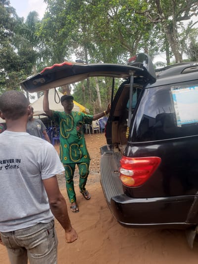 The image, taken in Umundugba, Nigeria, captures an outdoor scene featuring two men and a black SUV with its tailgate open.
In the mid-ground, a man wearing a vibrant green patterned traditional African outfit (Ankara print) and a black cap stands beside the open tailgate of the black SUV. He appears to be attending to the vehicle's trunk, possibly loading or unloading items. His hands are visible near the open tailgate. Behind him, several blue and white plastic chairs are stacked, suggesting a gathering or event is either in progress or being set up.
In the foreground, another man is seen from behind, wearing a grey t-shirt and grey jeans. Part of the text on his t-shirt is visible and reads "REVER IN R Hears," likely "FOREVER IN OUR Hearts."
The black SUV's rear is prominent, showing its red and orange taillights and a partially visible, inverted license plate with the number "060044163116/00" and some blue and white markings. The tailgate is propped open.
The setting is outdoors, characterized by a reddish-brown dirt ground and abundant green foliage, including tall trees with dense canopies in the background, suggesting a tropical or semi-tropical environment. The bright natural light indicates it is daytime. The overall impression is that of a casual outdoor activity, possibly related to a local event or daily life.
