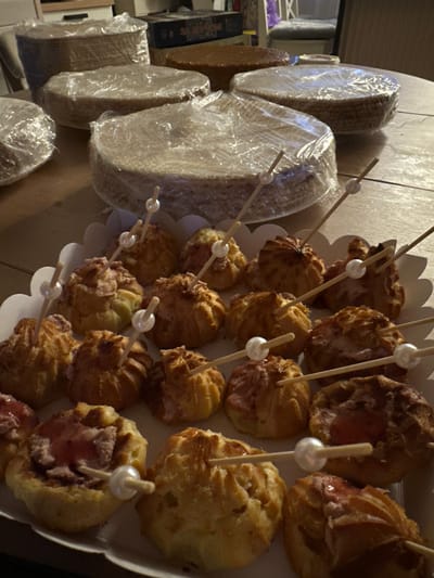 A close-up, high-angle shot shows a table laden with baked goods. In the foreground, a white, scalloped-edge tray holds numerous small, round pastries filled with a pinkish cream, each pierced with a wooden skewer topped with a pearl bead. Behind this tray, several large, round cake layers are stacked and wrapped in clear plastic. To the left and right of these cake layers, more plastic-wrapped cake layers are visible. Further back, a light brown cake with a smooth surface sits on the table. The setting appears to be indoors, with a wooden table surface and a hint of furniture in the background. The image captures a preparation scene for a bakery event or celebration.