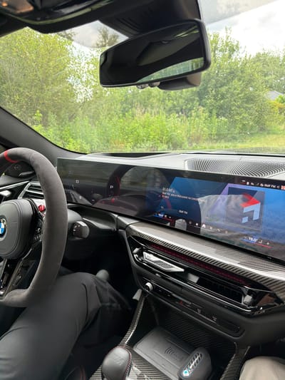 The image captures the interior of a car, likely a BMW, from the perspective of the driver. A prominent steering wheel with red stitching and a BMW logo is visible on the left. The dashboard features a large, curved digital display showing various information, including radio station "RMF FM" tuned to 91.0 MHz, playing in Warsaw on a frequency of 91 MHz. The current temperature inside and outside the car appears to be 19.0°C. The car's interior is finished with carbon fiber trim. The driver's legs, clad in dark trousers, are visible in the lower left portion of the frame. Outside the car, through the windshield, lush green foliage is seen, suggesting a location with abundant vegetation, potentially in or near the city of Dobrzyń, Poland, as indicated. The sky is overcast, with a hint of sunlight filtering through the trees. A rear-view mirror is mounted on the windshield.