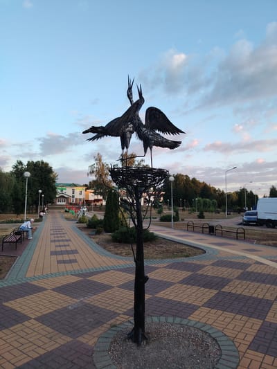 The image depicts a park or public square in Malaryta, Belarus. In the foreground, a metal sculpture of two cranes in a nest atop a tree stands prominently. The sculpture is dark, made of metal, with intricate details on the cranes' feathers and wings. The tree base is surrounded by gravel and a curb. A paved walkway with a checkered pattern of yellow, brown, and gray bricks curves through the scene.
To the left of the walkway, several people are present. One individual is seated on a park bench, facing away from the camera. Further down the walkway, more people are visible, including some walking and others sitting on benches. Adjacent to the walkway is a grassy area with trees and streetlights.
In the background, a light-colored building with a green roof is visible, possibly a public institution or community center. In front of the building, several flagpoles with flags are planted, and two silver statues of figures in uniform stand near the walkway. The sky above is a soft blue with scattered clouds, indicating either early morning or late afternoon light. A white van is parked on the right side of the image, alongside a grey car. The overall scene suggests a peaceful public space in the Belarusian town of Malaryta.