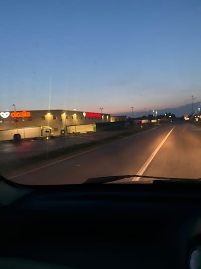The image depicts a road scene at dusk, with the sky showing gradient hues of blue and orange. The perspective is from inside a vehicle, looking forward through the windshield. The road ahead is paved and has a white line marking its center, illuminated by the headlights of the vehicle. On the left side of the road, a large building with illuminated signage is visible. The signage on the building reads "cicala" in red letters with a white logo. Further down the road, other buildings and streetlights are present. The ground appears wet, suggesting it may have recently rained. The scene appears to be in an urban or semi-urban environment, possibly an industrial or commercial area. The location is identified as Ponteterra, Italy.