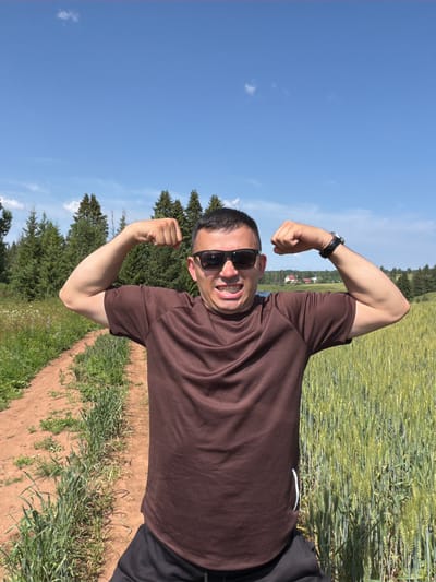 A man stands in a field, flexing his biceps. He is wearing a brown t-shirt and black shorts, and sunglasses. He has dark hair and is smiling. Behind him, a dirt path leads through a field of tall green grass and trees. The sky is blue with a few white clouds. The image was taken in Podshivalovo, Russia.