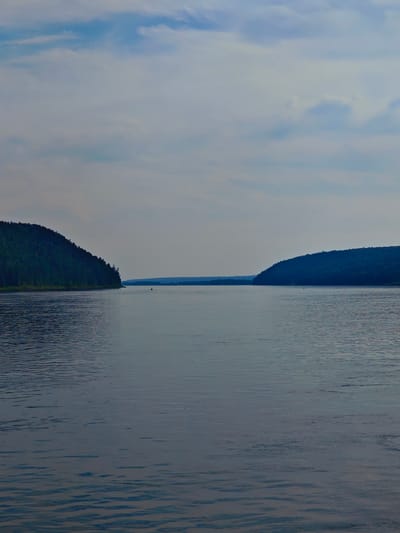 The image depicts a wide, calm river with a person in a small boat visible in the distance. On the left side of the river, a densely forested hill slopes down to the water. On the right side, a similar, less vegetated hill forms a concave shoreline. The sky is overcast with light clouds. This scene is likely the Angara River in Predivinsk, Russia, a region known for its vast waterways and forested landscapes.