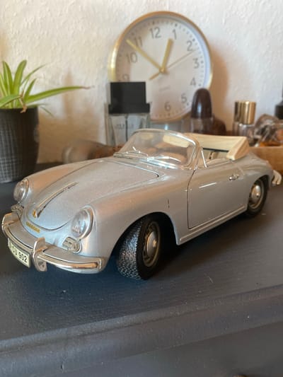 This image shows a close-up, low-angle, eye-level shot of a silver toy Porsche convertible. The car is parked on a dark, textured surface, possibly a shelf or table. A license plate on the front of the car reads "S-CU 902". In the background, a white wall with a textured finish is visible. To the left of the car, a small green plant in a dark pot is present. Behind the car, a round clock with gold hands and numbers is mounted on the wall, and various small bottles and objects are arranged. The lighting suggests an indoor environment, likely a domestic setting. The image does not contain any information that would allow for its location to be identified as Lütjenwestedt, Germany.