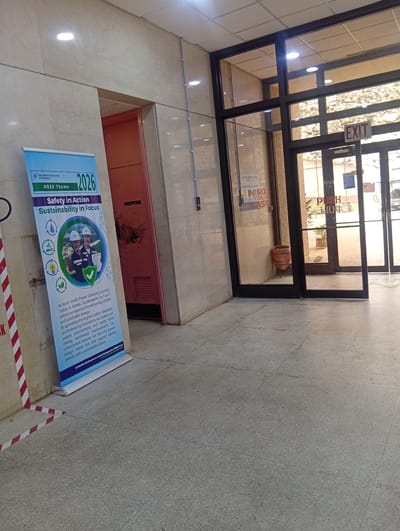 This is a full shot of an indoor hallway in Shiroro, Nigeria, likely at the North South Power Company. In the foreground, a blue and white banner stands angled to the left, displaying "NORTH SOUTH POWER" and "2026 HESS Theme: Safety in Action, Sustainability in Focus". Below this text is a circular image featuring two individuals in hard hats and safety vests, with a green checkmark emblem. Several circular icons representing water, recycling, and electricity are positioned to the left of this image. The banner continues with text detailing the company's commitment to safety and sustainability. The floor is tiled in a speckled grey pattern, and red and white caution tape is visible near the bottom left corner. To the right of the banner is a salmon-colored door with a ventilation grate and signage that is partially obscured. Further to the right, a glass-paneled entrance with black framing leads outside. A sign reading "EXIT" is visible above the entrance, and a "PUSH" sign is visible on the glass doors. Inside the entrance, a potted plant and a decorative orange pot are present. The overall environment appears to be well-lit with fluorescent ceiling lights.