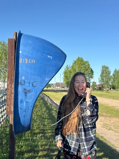 An outdoor scene in Ust'-Silayka, Russia, depicts an adult female interacting with a public payphone on a sunny day. The central subject is a bright blue, curved metal or plastic structure, resembling a partial cylindrical shell, mounted on a weathered brown square metal post. Yellow numbers "213111" are stenciled near the top of the blue structure, with darker, scribbled graffiti below, including characters that appear to read "G+C=" and a heart shape.

To the right of the payphone structure, a woman with long, light brown hair is shown from the chest up, holding a black, old-fashioned telephone receiver to her right ear. Her mouth is open wide in a joyful or excited expression, and her eyes are partially closed. She is wearing a dark blue and green plaid flannel shirt over dark pants patterned with red chili peppers. A light-colored wristwatch is visible on her left wrist.

The ground around the payphone and extending into the background is covered with green grass. A dirt road or path curves into the distance to the right. Sparse, light-green deciduous trees are visible throughout the background, and several small, rustic wooden houses are scattered in the mid-ground, indicating a rural village setting. A light-colored slatted fence is partially visible to the left of the payphone post. The sky is clear and bright blue, suggesting fair weather. The scene is illuminated by natural sunlight, casting distinct shadows.