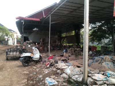 The image depicts an outdoor area in Gampongbaro, Indonesia, characterized by disarray and clutter. A white scooter is parked on a dirt ground, with a trailer attached behind it. The background features a large, open-sided structure with a corrugated metal roof, supported by metal beams. This structure appears to be a storage or workshop area, as it contains numerous piles of cardboard boxes, some stacked neatly and others scattered haphazardly. Among the boxes, there are also plastic bottles and other debris, indicating a state of neglect or a temporary storage situation for recyclable materials. The ground is covered in dirt and scattered trash, including plastic wrappers and bottles. In the distance, a building with a red roof and a white facade is visible, along with trees and foliage, suggesting a semi-rural or suburban setting. The overall impression is one of an unkempt, possibly industrial or recycling yard, with a mix of functional items like the scooter and trailer, and large quantities of discarded materials. The lighting suggests it is daytime, possibly overcast.