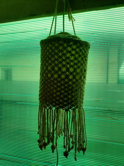 A macrame plant hanger with a cylindrical woven outer shell and a lighter green inner liner hangs against a translucent green corrugated plastic wall. The hanger is suspended by three strands of jute twine. The outer shell has a diamond-shaped pattern. The bottom of the hanger features a fringe of jute cords, some of which are adorned with small, dark wooden beads. The overall color palette is dominated by shades of green. The background wall suggests an indoor or covered outdoor space, possibly a greenhouse or porch. The location is stated to be Bolshoye Sorokino, Russia.