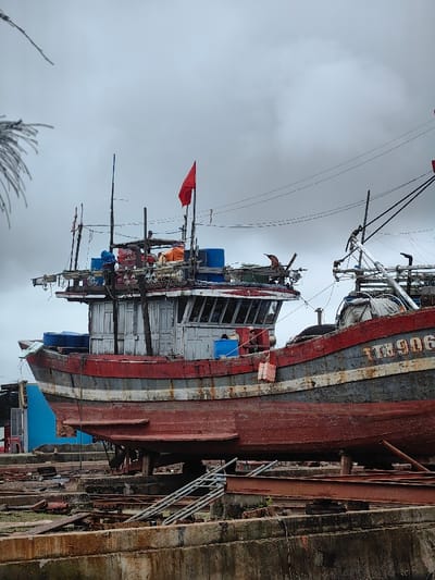 An weathered fishing vessel is dry-docked for maintenance in Thuận An Ward, Vietnam, under an overcast sky. The boat's hull is primarily reddish-brown with significant rust, a grey/black band, and a white stripe running along its length. The registration number "TTB 906" is visible on the bow section. A grey and white pilothouse with multiple windows sits atop the forward deck. A red flag, possibly the national flag of Vietnam, is mounted on a mast above the pilothouse. Various fishing equipment, including nets, ropes, and blue barrels, are stored on the deck. A person in an orange garment is discernible near the pilothouse on the upper deck. The boat is supported by concrete blocks and wooden shoring. The ground beneath and around the vessel consists of a dry dock area with rusty metal beams, debris, and a metal ladder or ramp. In the background, power lines traverse the cloudy sky, and a section of a blue structure is visible to the left of the boat's bow. Faint foliage is visible in the upper left corner.