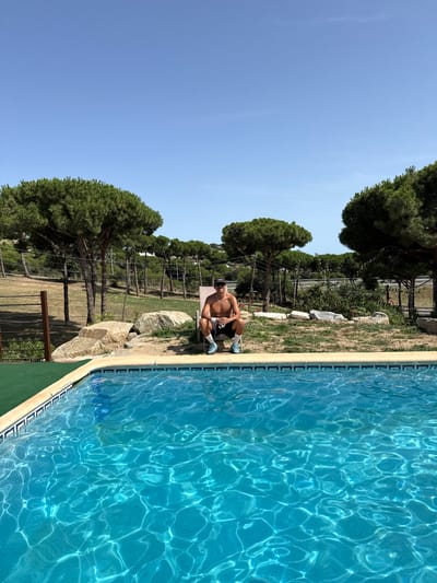 A young man, shirtless and wearing black shorts and a baseball cap, squats on a lounge chair beside a swimming pool. The pool water is a vibrant blue, reflecting the bright sunlight. Behind the pool, a grassy slope is dotted with rocks and mature pine trees. In the background, a highway can be seen, suggesting the location is near Sant Vicenç de Montalt, Spain, a town known for its proximity to coastal roads and natural landscapes. The sky is clear and a deep blue.