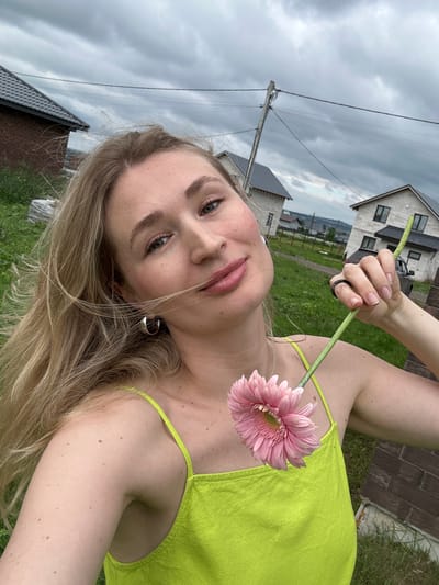 A young woman with blonde hair, wearing a lime green top, is holding a pink Gerbera daisy in her right hand. She is in a grassy outdoor area with several houses visible in the background. The sky is overcast. The location is the city of Mokino, Russia.