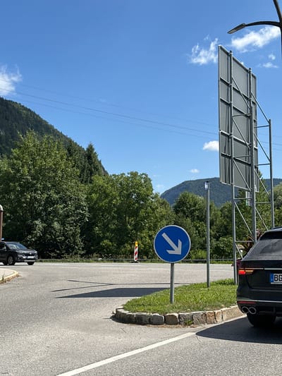 The image shows a road intersection in Berchtesgaden, Germany, on a sunny day. A black SUV is parked on the right side of the road, partially obscuring a large, gray advertising billboard supported by a metal structure. In the center of the image, a blue circular traffic sign with a white arrow pointing diagonally down and to the left is mounted on a pole. A black car is driving on the road to the left. The background features lush green trees and rolling hills under a clear blue sky with a few scattered clouds. The road is paved asphalt with white lane markings.