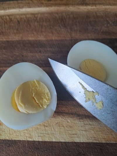 The scene displays two halves of a hard-boiled egg and a knife blade resting on a wooden cutting board. One egg half is positioned in the lower-left quadrant, showing a pale yellow yolk within a white albumen. The second egg half is in the upper-right, similarly presenting a pale yellow yolk. A silver-colored knife blade, with a pointed tip, lies diagonally across the mid-right section of the frame. The blade surface exhibits irregular fragments of pale yellow material consistent with egg yolk. The cutting board features a light-brown wood grain pattern with visible cut marks and scratches. The background is out of focus.
