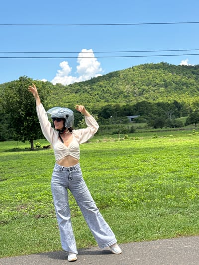 A woman wearing a light-colored, ruched crop top, acid-wash wide-leg jeans, and white sneakers stands on an asphalt path. She has on a silver motorcycle helmet with the visor up, sunglasses, and a red and white bandana tied around her neck. Her left arm is raised with her hand open, palm facing outward, and her right arm is bent at the elbow, with her hand held near her chest. She is positioned in front of a vibrant green, grassy field. In the background, a dense, lush green forest covers a hillside under a bright blue sky with scattered white clouds. Power lines are visible across the top of the frame, and a solitary tree stands to the left of the woman. The scene appears to be in a rural or semi-rural area, potentially near the city of Ham Thuan Bac, Vietnam, given the tropical vegetation and terrain.