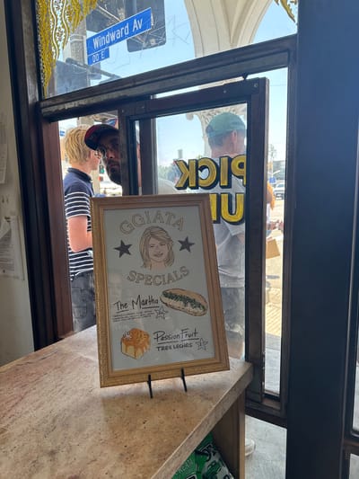 A medium shot shows a street-level storefront in Marina del Rey, California, United States. Through a open glass window of the storefront, a man wearing a green baseball cap and a striped shirt is visible. To his left, another man with blonde hair and a striped shirt is also seen. In the foreground, a framed sign displays a drawing of a woman with blonde hair and text that reads "GGIATA SPECIALS," "The Martha," and "Passion Fruit TRES LECHES." The sign also shows illustrations of a pastry and a sandwich. The storefront's interior features a marble countertop. Outside the window, a "Windward Av" street sign is visible.