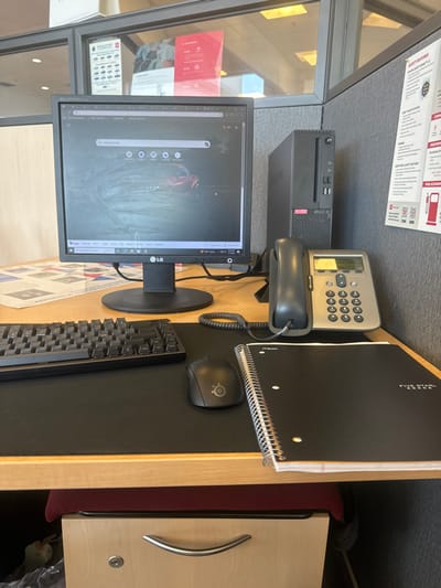 This image depicts an unoccupied workstation within a Toyota dealership in Newark, United States. The central element is a light wood-colored desk equipped with a black LG FLATRON computer monitor, displaying a smudged screen with a web browser showing a red car on a dirt background. To the right of the monitor stands a black Lenovo desktop computer tower. A grey digital desk phone with a coiled handset cord is positioned to the right of the computer tower. The desk surface features a black mouse pad supporting a black mechanical keyboard on the left and a black SteelSeries computer mouse to the right. A black Five Star spiral notebook lies open on the right side of the mouse pad. Below the desk, a light wood-toned two-drawer filing cabinet with a curved metal handle is partially visible, behind which a red chair seat can be seen. A white plastic bag is located to the left of the filing cabinet. The workstation is enclosed by office partitions: a grey fabric partition on the right displays a white poster detailing "TOYOTA SAFETY SENSE™ 2.5+" and "FUEL ECONOMY" with a gas pump icon. A glass partition on the left reveals additional red and white Toyota promotional posters, including one mentioning "LEASE OPTIONS," from an adjacent area. No individuals are present in the scene.