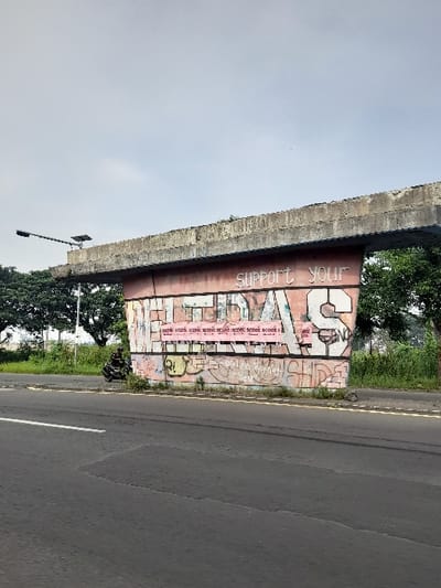The image displays a concrete overpass or bridge spanning over a grey asphalt road during daylight hours under a clear sky. The underside of the concrete structure is covered extensively in graffiti.
Prominently featured in white, bold, sans-serif lettering is the phrase "SUPPORT YOUR-", with the subsequent word obscured or incomplete. Below this, a dense layer of colorful and stylized graffiti covers the surface. A notable element is a pink rectangular banner-like section containing smaller white text, parts of which are legible. This text appears to be a political message, with discernible phrases such as "KAMI RAKYAT INDONESIA" (We the people of Indonesia) repeated several times. Other visible words include "DEMOKRASI" (Democracy), "KEADILAN" (Justice), "TOLAK" (Reject), and "HUKUM" (Law), indicating a protest or call to action. Large, stylized graffiti letters, possibly spelling "SAVE US FOR", are also integrated into the artwork.
Beneath the bridge structure, to the left, there is green vegetation, including trees, and a street light pole equipped with a solar panel. The asphalt road itself shows a faint white line towards its right edge. No people or active events are present in the scene, which appears to be in an urban or semi-urban area of Kecamatan Gempol, Indonesia.