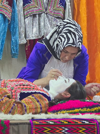 Two people are in the frame, oriented vertically. The person on the left wears a black and white zebra-patterned headscarf and a bright blue robe. They appear to be lying down or reclining, with their eyes closed. A white lace garment is visible underneath the blue robe.
The person on the right is applying a white mask to their face with a small applicator. They have dark hair pulled back and are wearing a colorful, embroidered vest or jacket over a purple garment. The vest features intricate beadwork and embroidery in shades of pink, orange, yellow, and purple. Their expression is neutral as they focus on applying the mask.
Surrounding the subjects are various textiles. To the left, there are several pieces of clothing or fabric covered in shimmering silver and blue sequins, with colorful embroidered patterns. To the right, there are more vibrantly colored textiles with fringe and geometric patterns, including a section of pink, yellow, and green fringe and a patterned textile with zigzags in pink, yellow, green, and black. An orange fabric serves as a backdrop behind the subjects. The scene appears to be indoors, possibly in a dressing room or a space set up for preparing for an event. The overall impression is one of preparation and cultural display, with an emphasis on traditional or ceremonial attire. There is no visible text in the image.