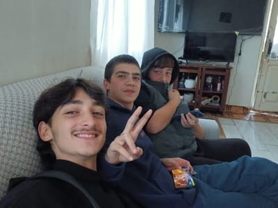 Three young males are depicted seated on a textured, light-colored sofa indoors in the city of ქვედა სამება, Georgia. The individual in the foreground, positioned to the left, has dark hair, a mustache, and a goatee, smiling towards the camera while making a peace sign with his right hand. He is wearing a dark long-sleeved top and has a smartphone displaying a colorful screen resting on his lap. To his right sits a second male with short dark hair, wearing a dark blue long-sleeved top, looking directly forward. Partially obscured behind him on the right is a third male, wearing a dark hooded vest with the hood up, holding a dark smartphone in his right hand. The background reveals a light-colored wall and a window with white curtains on the left. To the right, a dark flat-screen television is mounted on a wall above a wooden shelving unit containing various bottles and objects. Below the wall-mounted television, another smaller screen is visible on the shelving unit. The floor is tiled with a brown and beige geometric pattern. A light-colored doorframe or door is visible at the far right.