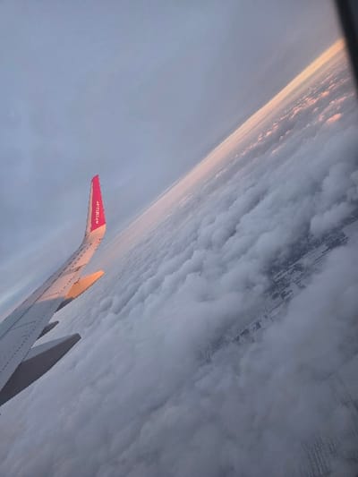 The image displays an aerial view captured from an aircraft window, angled upwards and to the right. A portion of a light gray airplane wing is visible in the lower-left quadrant, featuring a bright pink winglet with "wizzair.com" written vertically in white text. The wing's upper surface is illuminated by a soft, warm light. Below and to the right of the wing, a vast, dense layer of white and gray cumulus clouds extends, partially obscuring the ground below. Through intermittent breaks in the cloud cover in the lower right, patterned segments of land, suggestive of agricultural fields or infrastructure, are discernible. The upper right portion of the image shows a clear horizon line, marked by a band of golden-pink light against a pale blue-gray sky, indicative of either sunrise or sunset. The aircraft is situated over or near the city of Yagel, Israel. No individuals are visible in the frame.