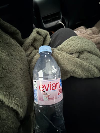 An Evian water bottle with a blue cap is placed on a dark surface, likely a car seat or floor, surrounded by plush, olive-green and beige blankets. The water bottle's label is visible, featuring the Evian logo in pink and an image of a tennis player. The background is dark, suggesting an indoor environment such as a car's interior, with a hint of a car's air vent visible in the upper right corner. There are no people present in the image. The location is unspecified but could be Holdorf, Germany, if the context suggests it.