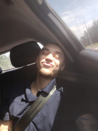 The image shows a young man sitting in the passenger seat of a car, with his eyes closed and a smile on his face. He is wearing a dark blue polo shirt and a silver chain necklace. A seatbelt is across his chest. Sunlight streams in through the car's sunroof, illuminating his face and casting shadows. Outside the car, trees and a street light are visible, suggesting the car is in a suburban or rural area, possibly on the outskirts of a city. Given the user's prompt, this scene could be occurring in Perovo, Russia, where such environments are common. The man appears relaxed and content, enjoying the ride.