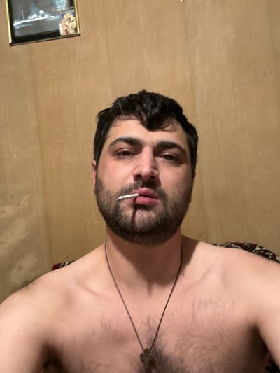 A man with dark hair and a beard holds a cigarette in his mouth, looking directly at the camera. He is shirtless, wearing a necklace. The background is a plain, textured wall, with a framed picture hanging on it in the upper left corner. The lighting appears to be indoor, with a warm tone. The scene does not provide specific information to determine its location within the city of Gyumri-Akhuryan, Armenia.