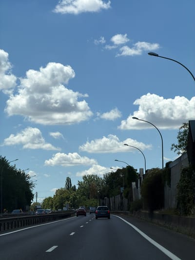 This is a vertical, outdoor, daytime photograph taken from a moving vehicle on a highway. The scene depicts a sunny day with scattered clouds.
The primary subjects are the highway, several cars traveling on it, and the sky filled with cumulus clouds. The highway appears to be a multi-lane road with a white line marking the lanes. Along the right side of the highway, there is a guardrail and then a row of trees and a concrete barrier. Several streetlights with curved arms are visible along the left side of the road, arching over the highway.
The activity taking place is normal highway traffic, with cars moving in the same direction. The overall impression is one of travel on a clear, pleasant day. The location cues suggest a developed area with infrastructure for transportation, consistent with a highway in Reckange-sur-Mess, Luxembourg. There is no visible text in the image.