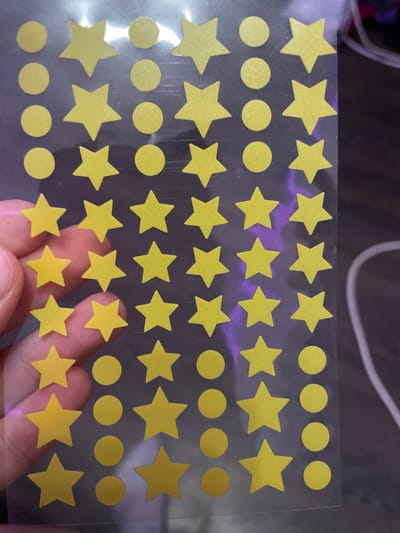 The image shows a sheet of yellow star and circle stickers. The stickers are arranged in a grid pattern on a transparent plastic sheet. The sheet is held by a person's hand, partially visible on the left side of the image. The background is dark and out of focus, with some purple and white light reflections. There is no indication of the location in Romanovka, Russia.