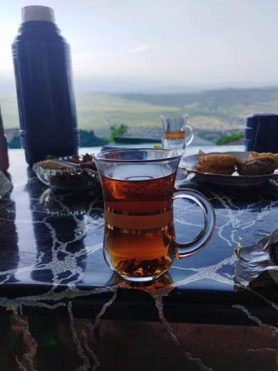 A close-up, eye-level shot focuses on a glass of amber-colored tea with gold bands, placed on a dark, reflective table with a textured surface. Behind the tea, a small glass cup also filled with tea sits with a thermos to its left. A plate of pastries is visible in the background, along with another dark blue cup. The scene appears to be outdoors, overlooking a hazy landscape with rolling hills and a town nestled in the valley below, suggesting a location near Darvag, Russia, given the prominent thermos often used for hot beverages in colder climates and the presence of pastries, suggesting a break or picnic. The sky is overcast, with soft light illuminating the distant scenery. There are no people visible in the frame.