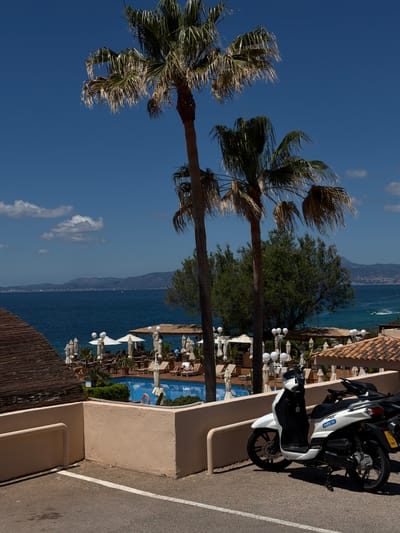 The image captures a vibrant scene in Llucmajor, Spain, likely near the coast. Two tall palm trees dominate the left side, their fronds swaying in the wind against a clear blue sky with scattered clouds. In the background, the deep blue sea stretches towards a distant mountain range.

A resort or hotel is visible on the right side, featuring a swimming pool surrounded by sun loungers, umbrellas, and other greenery. People can be seen enjoying the pool area. A row of globe-shaped lamps lines the edge of the resort, adding to the ambiance. A parking area is to the far right of the resort where a motorcycle is parked. A low wooden fence is partially visible in the bottom left corner, adding a rustic touch to the scene.