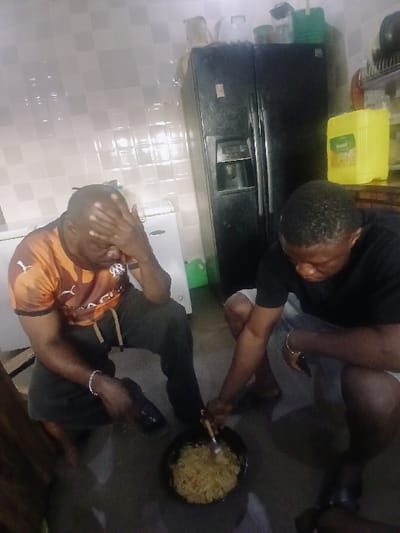 The scene depicts an indoor kitchen environment, specifically within Kungulu School, Zambia. Two adult males are observed in a crouching position. The individual on the left, wearing an orange and black short-sleeved shirt with a partial "MARSEILLE" logo, dark trousers, and a silver bracelet on his left wrist, rests his right hand on his forehead, looking downwards. A black shoe is positioned near his right foot. The man on the right, dressed in a black short-sleeved t-shirt and light-colored shorts, holds a metal utensil and actively stirs a light-colored, possibly noodle or rice-based dish contained within a black pot situated on the dark concrete floor. A gold-toned bracelet is visible on his right wrist. The background features light-colored square tiled walls. A dark-colored double-door refrigerator with a water dispenser is prominent on the right, next to a smaller white appliance. A large yellow jerry can, bearing the label "Grandi," is visible on a counter, with additional containers stacked atop the refrigerator.