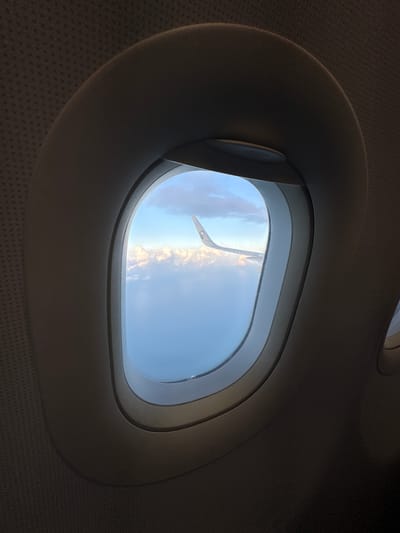 This image depicts a view from an airplane window, looking out at the sky and clouds, with a portion of the aircraft's wing visible. The interior of the airplane cabin is visible around the window frame. The scene is of an aerial perspective, likely above the clouds. There is no indication of a city, nor is there any specific visual information that could pinpoint the location to Bandar Penawar, Malaysia. The image is a standard travel photograph taken from a passenger aircraft in flight.