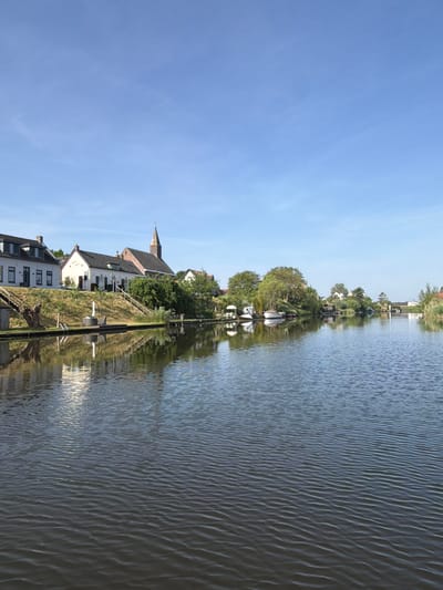 The image depicts a canal scene in Gellicum, Netherlands. Buildings with white facades and dark roofs, including a church with a spire, are visible along the canal bank. Several small boats are docked along the water's edge. The water surface shows ripples and reflections of the surrounding buildings and trees. The sky is blue with a few scattered clouds. The image is taken from a perspective parallel to the canal, presenting a clear view of the canal and the adjacent infrastructure.