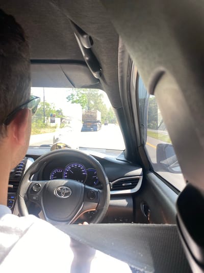 The image shows the interior of a car, specifically the driver's perspective. The driver, a man, is visible in the bottom left corner. He is wearing glasses. The dashboard and steering wheel, bearing a Toyota logo, are prominent in the center. Outside the windshield, a street scene is visible with a truck and some signage. The photograph was taken in the city of บ้านท่าฉัตรไชย, Thailand.