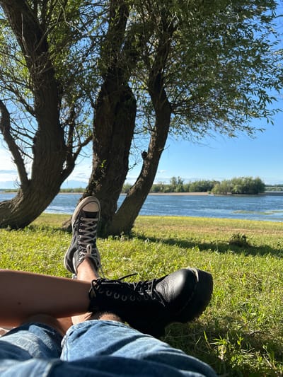 The image depicts a low-angle shot of two people sitting on a grassy area with their legs extended towards the camera. One person is wearing black Converse sneakers and denim shorts, while the other is wearing black Dr. Martens boots and denim shorts. In the background, there are large trees with rough bark and lush green leaves. Beyond the trees, a body of water, likely a river, is visible under a clear blue sky with scattered clouds. The opposite bank of the river features a line of trees and vegetation. The scene suggests a relaxed outdoor setting. The provided crops do not offer any specific information to confirm a location in Florentin, Bulgaria.