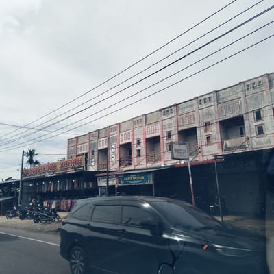 This image captures a daytime street scene in Teungoh Baro, Indonesia, under an overcast sky. The prominent subjects include a row of two-story concrete buildings lining the right side of the road, featuring multiple balconies and visible signs of wear with peeling paint. Overhead, numerous power lines crisscross the frame against the pale sky.
In the foreground, a dark-colored car, possibly an SUV or MPV, is captured in motion, its body blurred, suggesting movement. On the left side of the road, several motorcycles are parked near a shop. This shop, identified by a large yellow sign, displays an array of clothing on racks outside. A person is partially visible near these clothes racks.
Visible text on various signs provides commercial and service information:
* "SUNAN SERBA MURAH" with additional text "ARANG DENGAN HARGA DISTRIBUTOR JL. BANDA ACEH MEDAN - KEUDE CALEU Community" for the clothing shop.
* "BENGKEL ALIFA MOTOR" for a motor repair shop.
* "PRAKTEK GIGI" and "GIGI PALSU JASRINANDA, A. Md. KG" for a dental practice.
* A partially visible sign reads "BUMDESM AMAINDRAJ KECAMATA PRATAYA KABUPAT".
* Another small sign says "TEMPEL BAN".
The scene depicts a typical commercial street with various local businesses, suggesting an active urban or semi-urban environment.