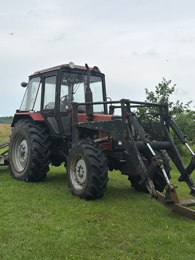 A dark red tractor with a front loader attachment sits on a grassy field under an overcast sky. The tractor is facing to the right of the frame, with its large rear tire prominently displayed. The front loader is painted black and appears weathered, with visible hydraulic lines and metal components. A small tree with green foliage is visible to the right of the tractor, and a distant line of trees can be seen on the horizon. Two birds are present in the sky, one on the left side and another on the right. The location is described as Ozoldangas, Latvia.