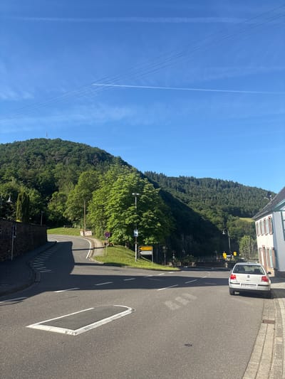 The image depicts a street scene in Schuld, Germany, on a clear day. The foreground shows a road with white lane markings and a speed bump. A white Volkswagen Golf with license plate AW H3200 is parked on the right side of the road. Adjacent to the car is a sidewalk made of stone pavers, and a building with white walls and green shutters is visible.

The road curves to the left, leading towards a densely forested hill that dominates the background. The hill is covered in green trees, with a radio tower visible at its summit. The sky is a vibrant blue with a few wispy clouds and contrails from an aircraft.

On the left side of the road, there is a stone wall and a lamppost. Further up the road, yellow street signs indicate directions and distances to locations such as "Nittenburg 4 km" and "Wissenbach 10 km," as well as "Freilichtbühne." A traffic sign with a red circle and a diagonal bar is also present. The overall environment suggests a rural or semi-rural setting, possibly on the outskirts of the town.
