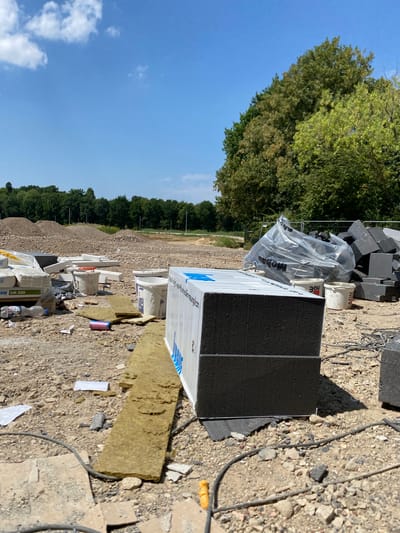 This image depicts a construction site under a clear blue sky with scattered clouds. In the foreground, there is a large, dark-colored rectangular object, possibly a building material, with white lettering on its side. To the left of this object, several white bags, possibly containing cement or mortar, are visible, one with the label "SCHAUER SM 300". There are also white buckets, some containing material, and a partially filled plastic water bottle. A long, yellowish-brown insulating material lies on the ground. In the middle ground, there are piles of construction debris, including gravel and broken concrete. To the right, a pile of dark gray blocks is stacked, and a large transparent plastic bag with the word "mobau" printed on it is visible, containing what appears to be more building materials. Further in the background, there are large mounds of dirt and a line of trees. The overall scene suggests ongoing construction or renovation work. The context provided indicates the location is in Hückelhoven, Germany.