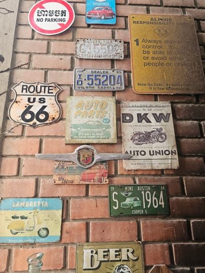 The image depicts a section of a brick wall adorned with a collection of vintage and decorative signs. These signs include an "Alpine Responsibility Code" plaque, a "No Parking" sign in both Cambodian script and English, license plates from Costa Rica (1972), North Carolina (1993), and a green "Mini Austin 1964" plate. Other signs showcase vintage advertisements for "DKW," "Auto Union," "Auto Parts," and "Lambretta." A Route 66 sign and a "Beer" sign are also present. An emblem with a shield design and the word "Ibiza" adds to the collection, as does a chrome car hood ornament. The overall impression is a decorative display, possibly on the exterior of a building, with a collection of vintage signs and automotive-related items. The location details, as provided, suggest it is in La Molina, Peru.