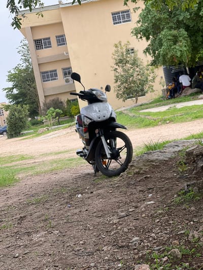 The image shows a motorcycle parked on a dirt path next to a light yellow building. The building has multiple windows with white frames. Green trees are visible in front of and to the side of the building. A group of people, some in white shirts, are gathered in the background near the building. The ground is a mix of dirt, small stones, and sparse green grass. The sky is overcast, suggesting a cloudy day. The motorcycle is white with black accents, including the wheels and handlebars. It appears to be a modern scooter-style motorcycle. There is no visible text in the image. The setting is outdoors in what appears to be a relatively rural or semi-urban area, given the dirt path and the building's design.
