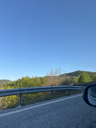 The image, taken from inside a vehicle, shows a road with a gray metal guardrail to the left. Beyond the guardrail is a tree-covered hillside in varying shades of green. Some trees are bare. A car's side mirror is visible in the upper right corner. The sky occupies the left half of the frame and is a clear blue with a contrail visible near the bottom. A piece of plastic litter lies on the side of the road. The location is on a road near Echinos, Greece.