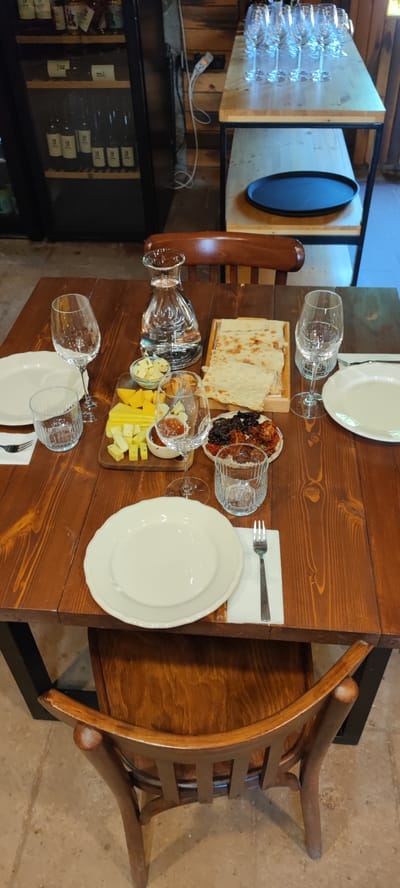 The image shows a close-up view of a table set for a meal, likely in Taperakan, Armenia. The table is made of dark wood and is set with white plates, wine glasses, and a carafe of water. There is also a wooden board with cheese and what appears to be dried fruits and nuts. Two chairs with wooden backs and seats are visible at the table. In the background, there is a wooden shelf unit with several bottles and glasses. The lighting suggests an indoor setting, possibly a restaurant or home.